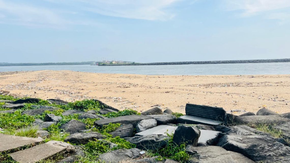 Person standing barefoot on the estuary shore at sunset, with gentle waves and scattered shells.